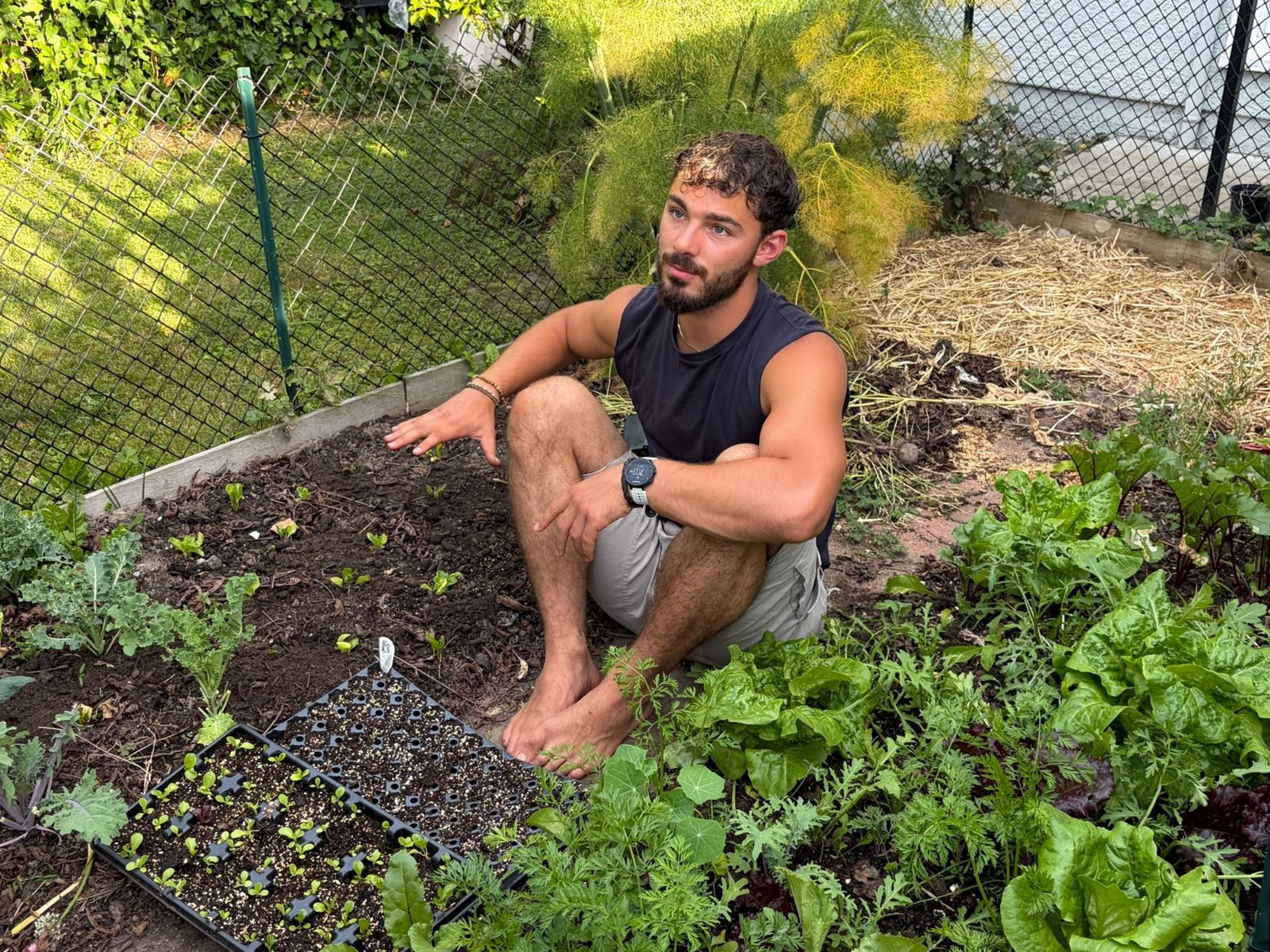 Nathan sitting barefoot in a garden bed among seedlings, greens and fennel