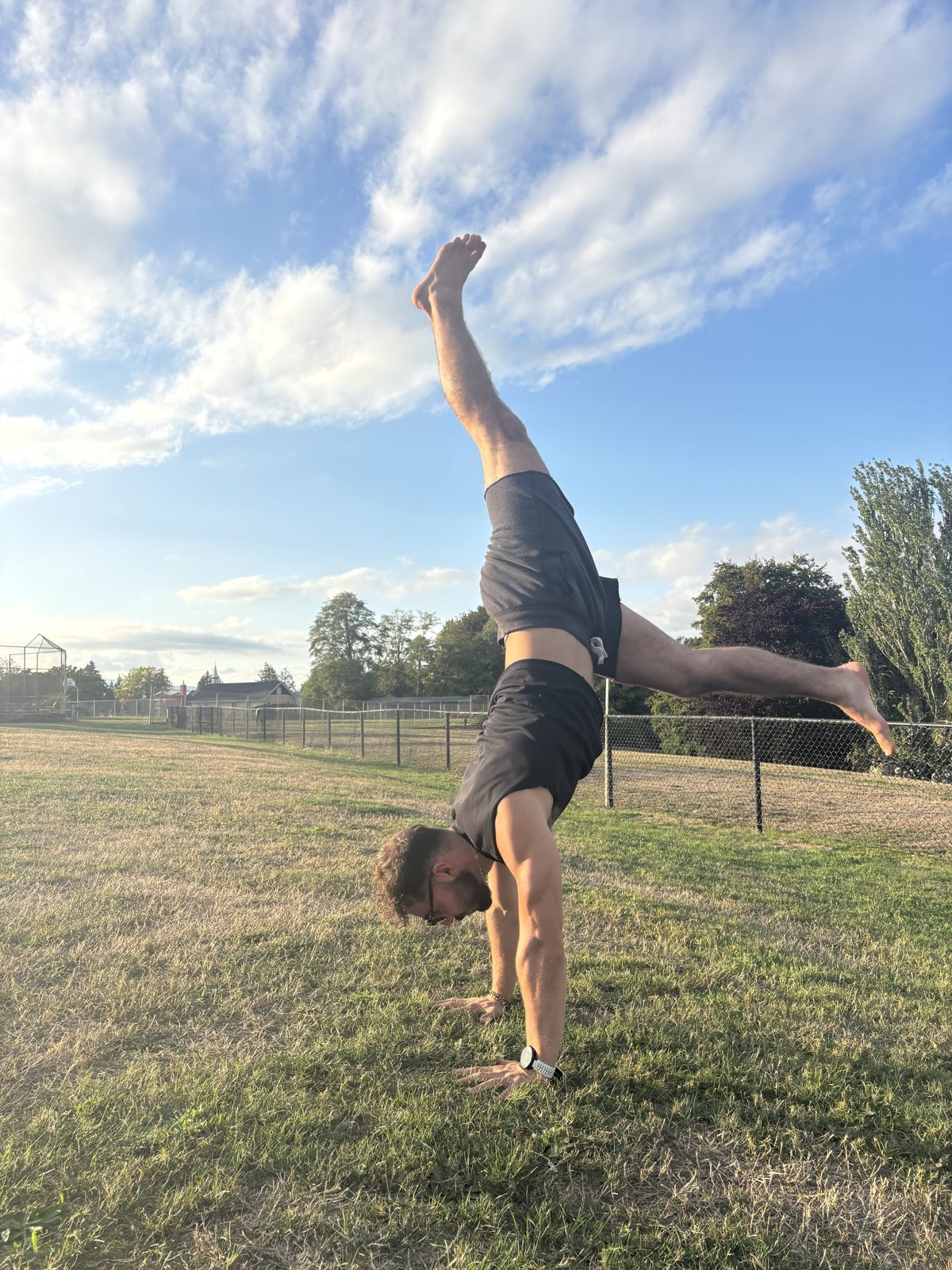 Nathan in a split-leg handstand on the grass of a sunny park under a wide blue sky