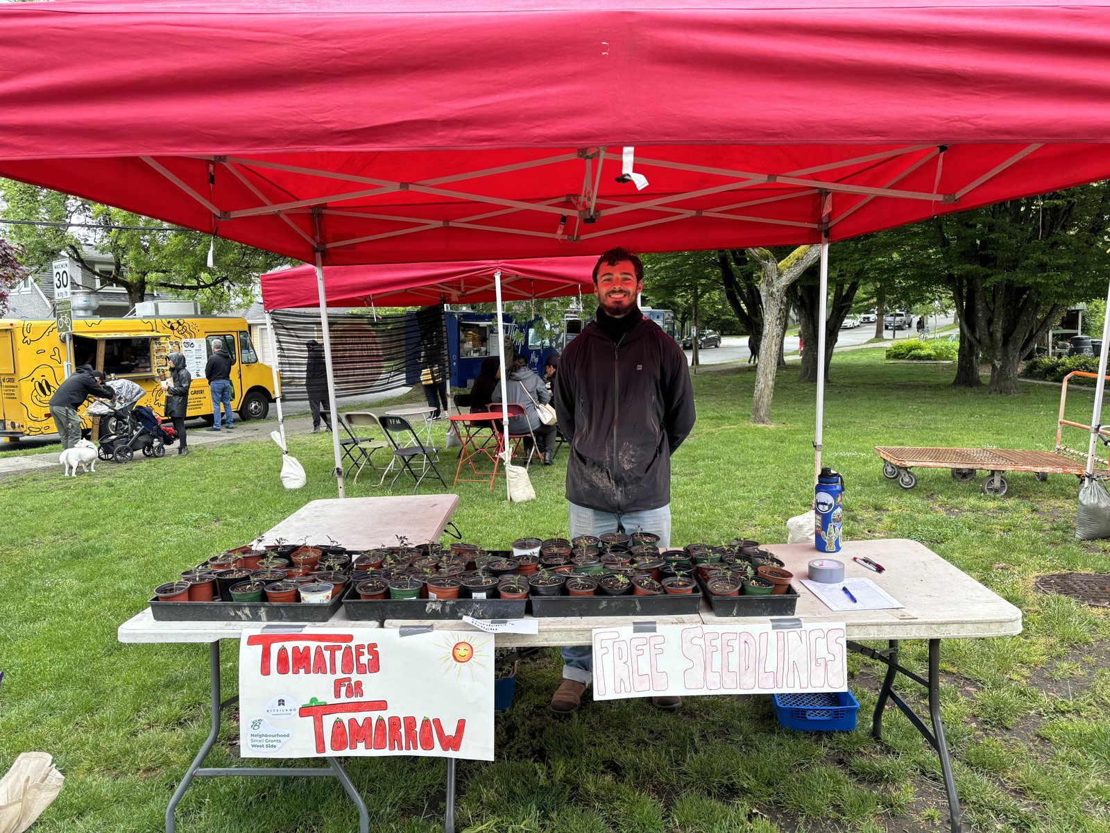 Nathan smiling under a red tent behind tables of free tomato seedlings